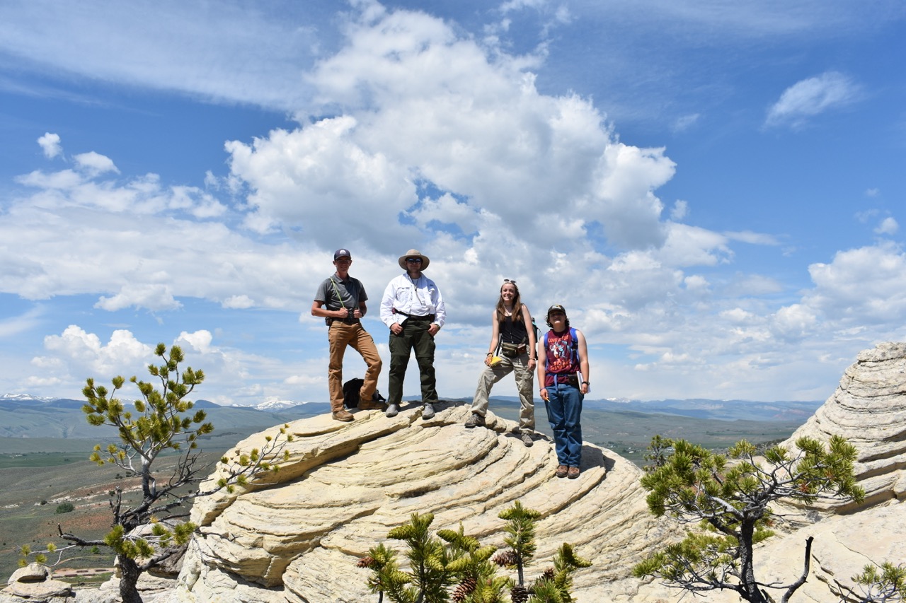 Four people stand on layered rock formations under a bright blue sky. The landscape features distant mountains and sparse greenery. The mood is adventurous and energetic.