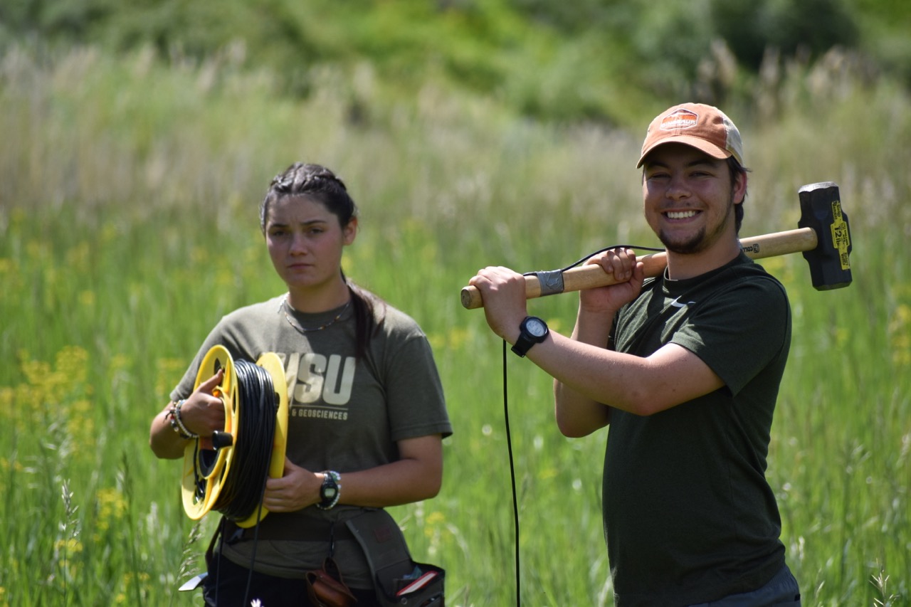 Two people stand in a grassy field. One holds a coiled cable, looking serious. The other smiles, resting a sledgehammer on their shoulder. The scene conveys outdoor work or research.
