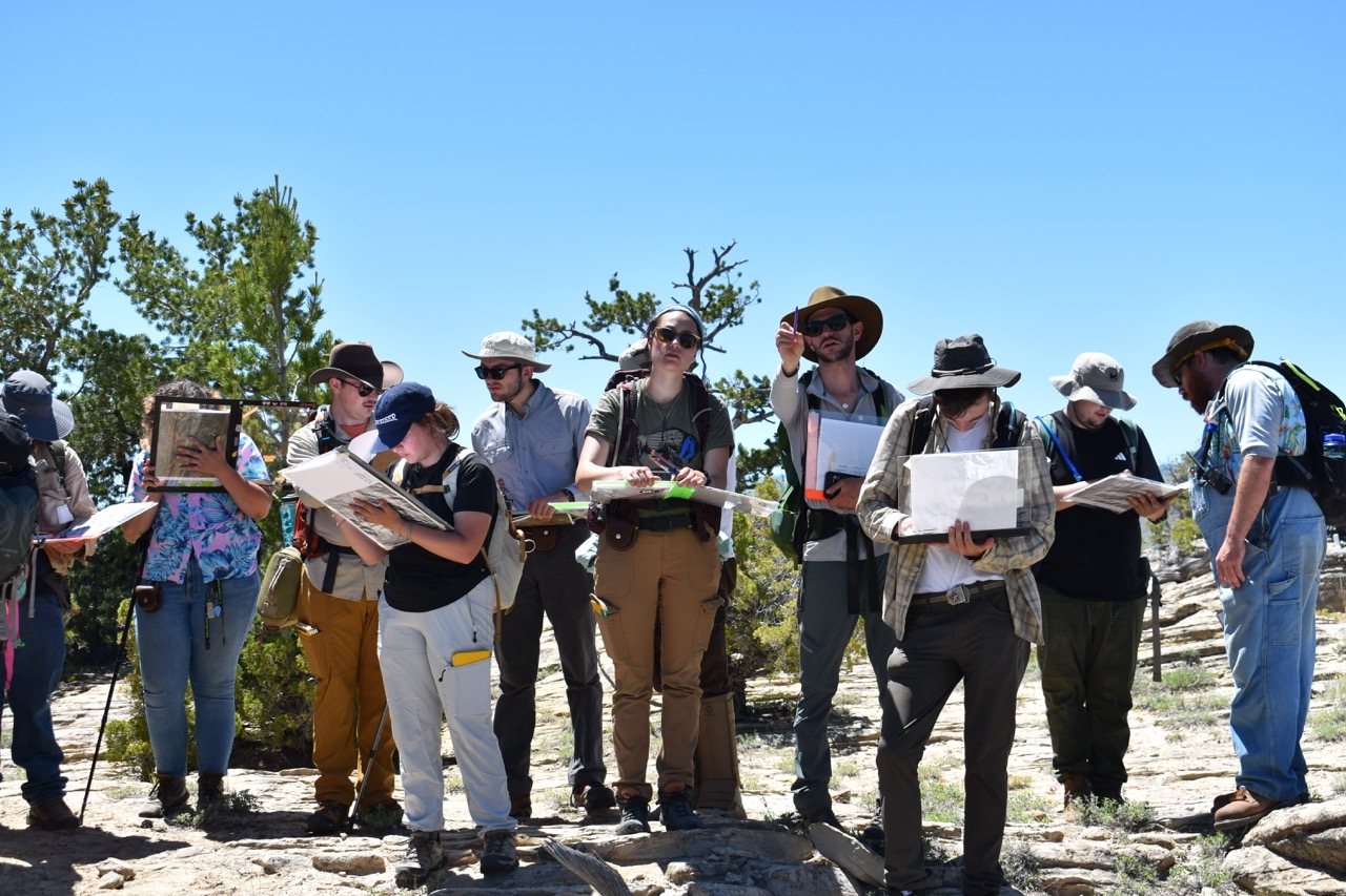 A group of people in outdoor gear, holding maps and compasses, stand amidst a rocky landscape with sparse trees under a clear blue sky. They appear focused and engaged.
