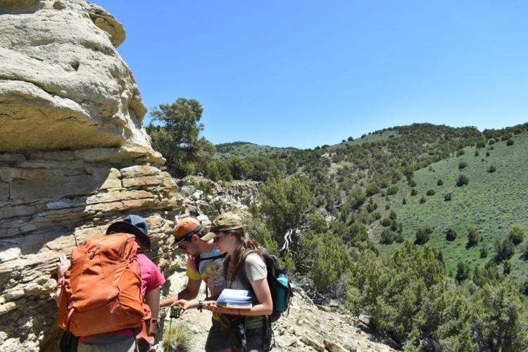 Three hikers examine a rock formation under clear blue skies in a hilly, green landscape. They appear curious and engaged, carrying backpacks and gear.