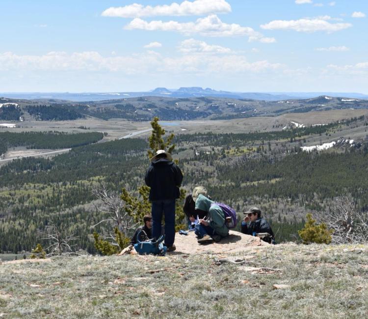 Four people rest on a grassy hilltop overlooking a vast landscape of lush forests and distant mountains under a partly cloudy sky.