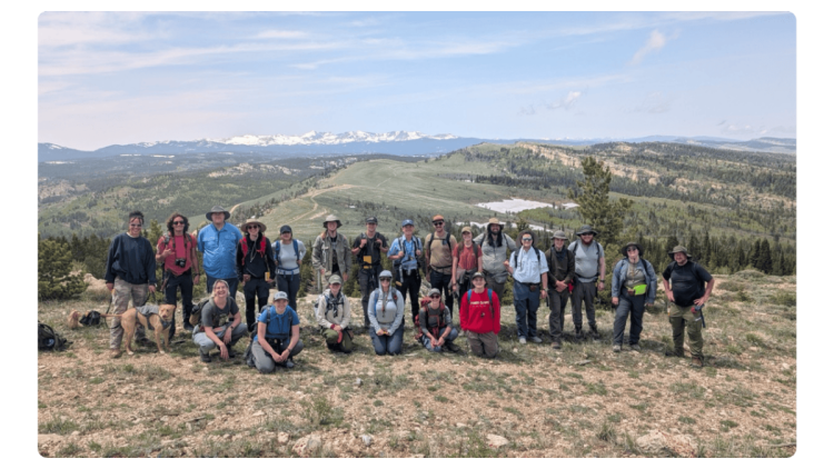 A diverse group of field camp students pose on a rocky hilltop under a bright sky, with green forests and distant snow-capped mountains in the background.