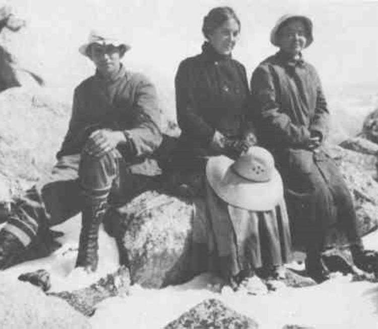 Three people sit on snowy rocks during a mountain expedition. They wear heavy clothing and hats, conveying a sense of adventure and endurance.